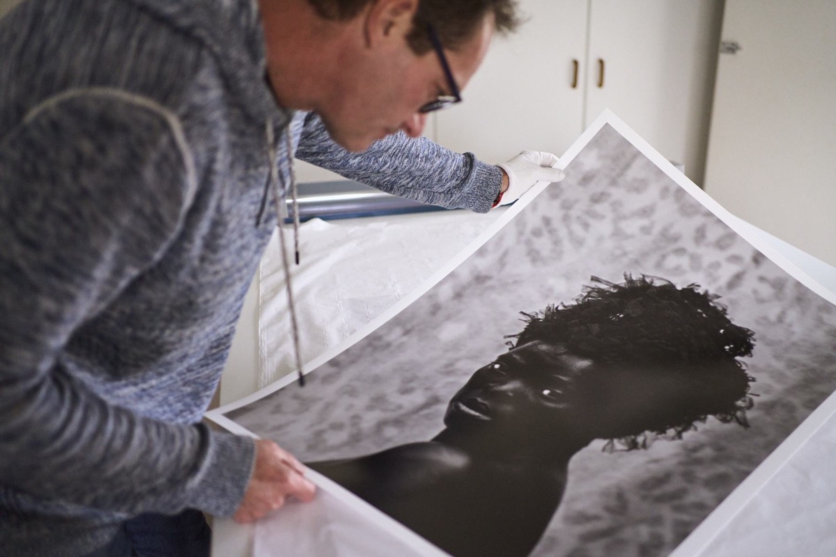 Vincent van Graan in white cotton gloves, bent over a large black and white portrait print on textured paper, checking it on the studio table