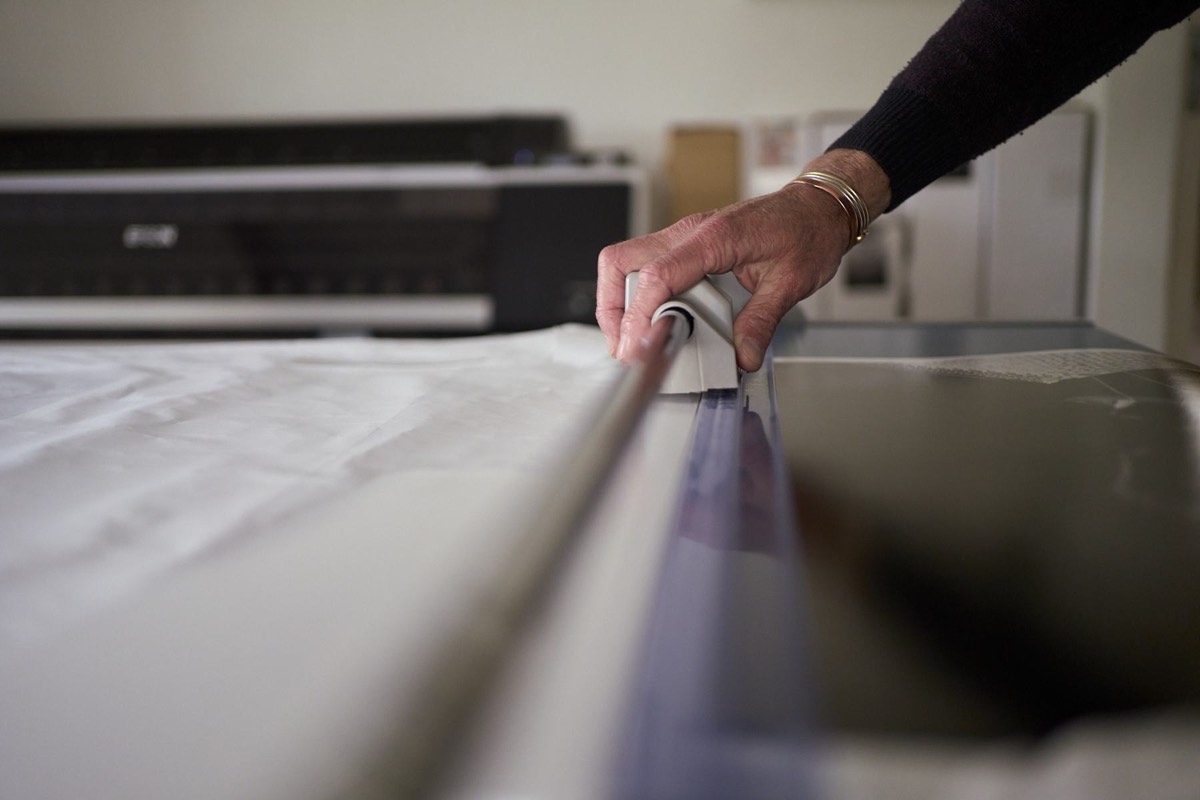 Close-up of a hand drawing a steel straight edge along the border of a print, the printer out of focus behind