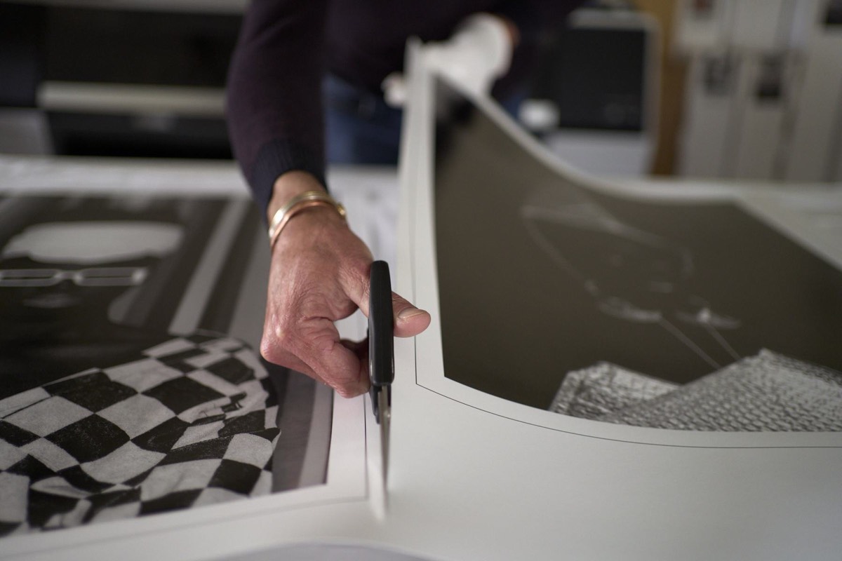 Close-up of a gloved hand cutting the edge of a print with a blade, a checkerboard patterned print beside it