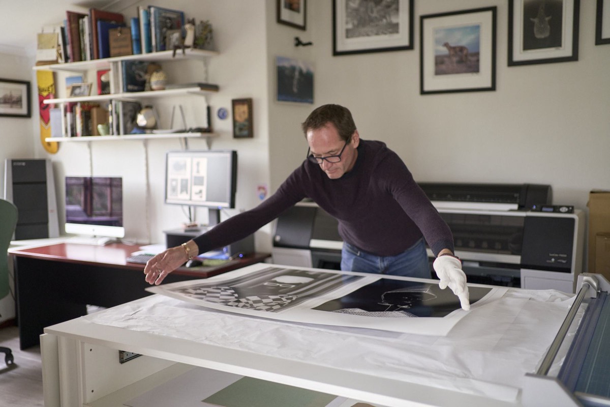 Wide view of the studio: Vincent reaching across the cutting table where several prints are laid out, framed photographs on the wall
